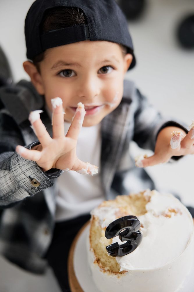 Smiling boy showing frosting-covered fingers over a half-eaten white cake with a black '3' candle