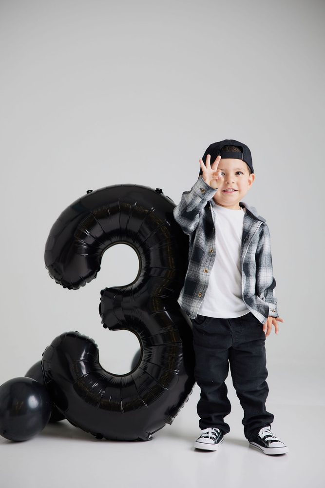 Boy in plaid shirt and black jeans making an OK sign beside a black number 3 balloon and black balloons
