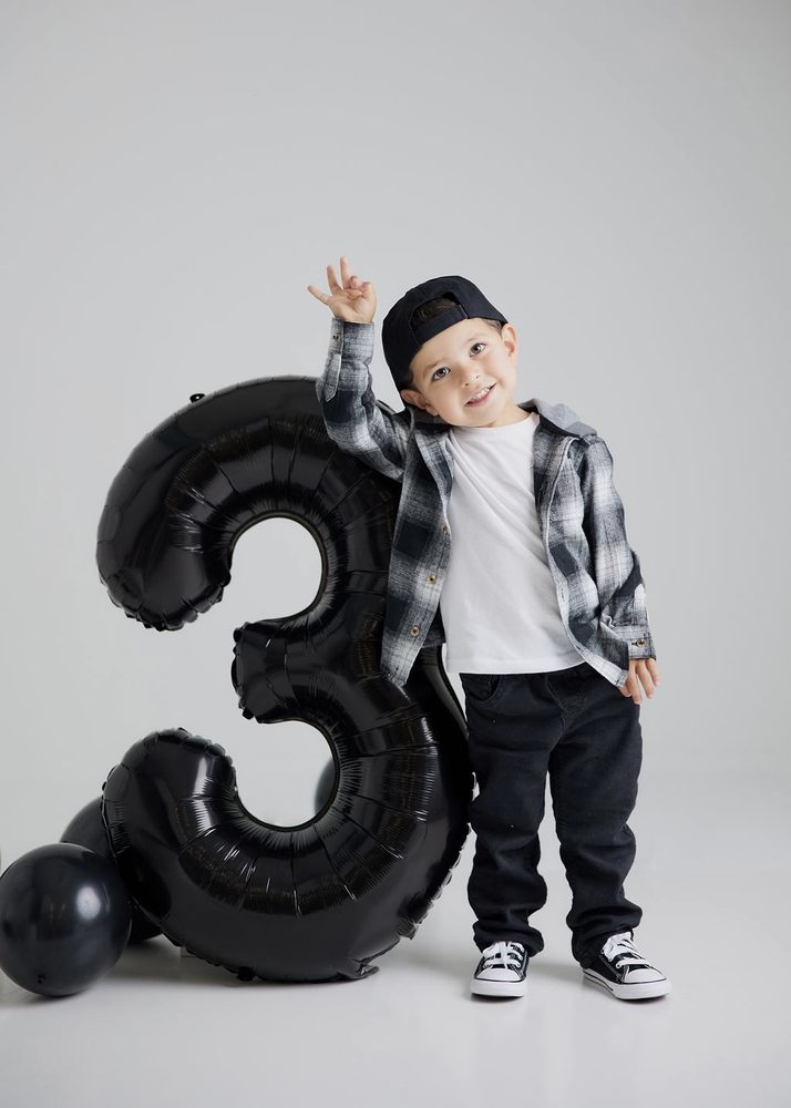 Boy waving with one hand raised while leaning on a giant black '3' balloon against a white studio backdrop