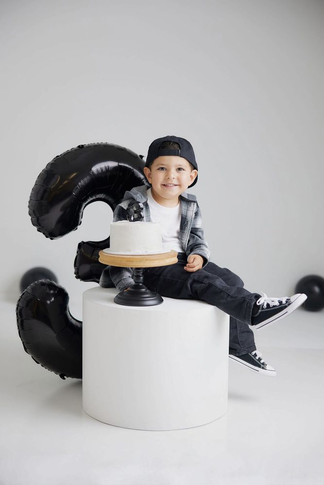 Boy sitting on a white pedestal next to a white cake on a stand, a black '3' balloon behind him