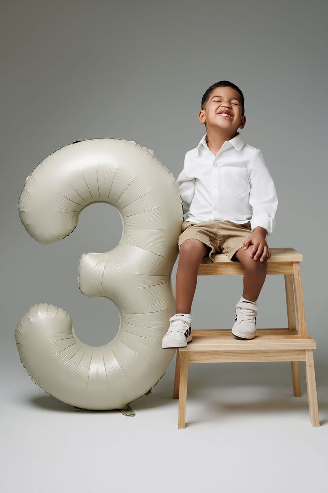 Boy squeezing his eyes shut in a big laugh, hands on knees on a step stool next to a satin '3' balloon