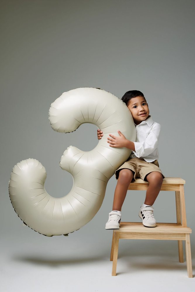 Boy in white shirt and khaki shorts hugging a cream '3' balloon while seated on a wooden step stool