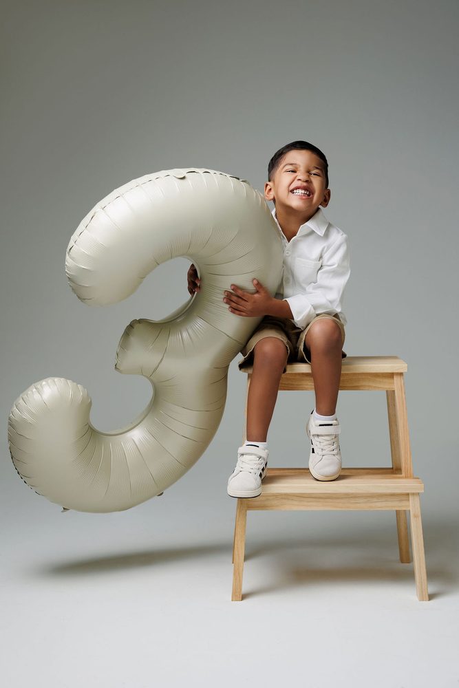 Grinning boy squeezing an oversized satin '3' balloon from his perch on a wooden step stool