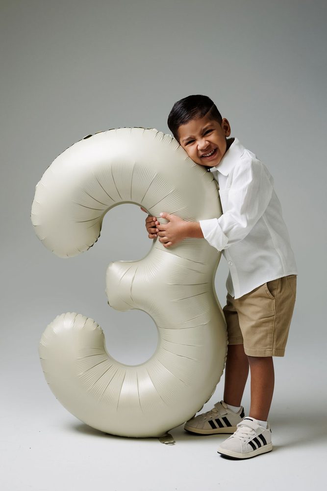 Boy laughing with his cheek pressed against a cream '3' balloon he hugs while standing on a gray backdrop