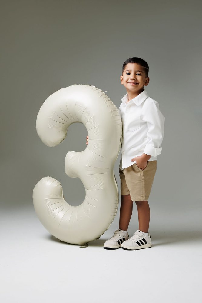 Boy in white button-down and khaki shorts standing with a hand in his pocket beside a cream '3' balloon