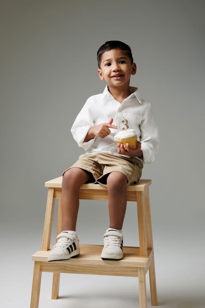 Boy on a wooden stool pointing at the frosted cupcake he holds, topped with a gold '3' candle