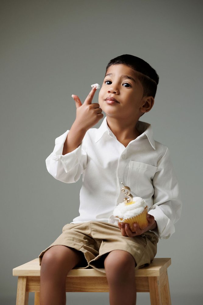 Boy licking frosting from his finger while holding a cupcake with a gold '3' candle