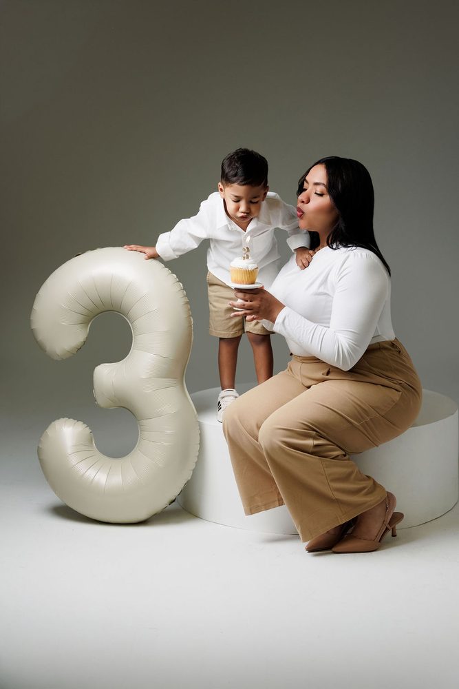 Mother and son blowing out a gold '3' candle on a cupcake beside a cream number balloon