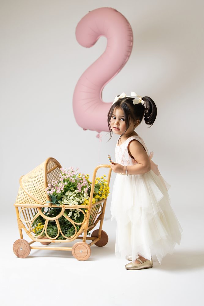 Toddler girl in a white tulle dress and hair bows beside a rattan doll pram of flowers, pink '2' balloon above