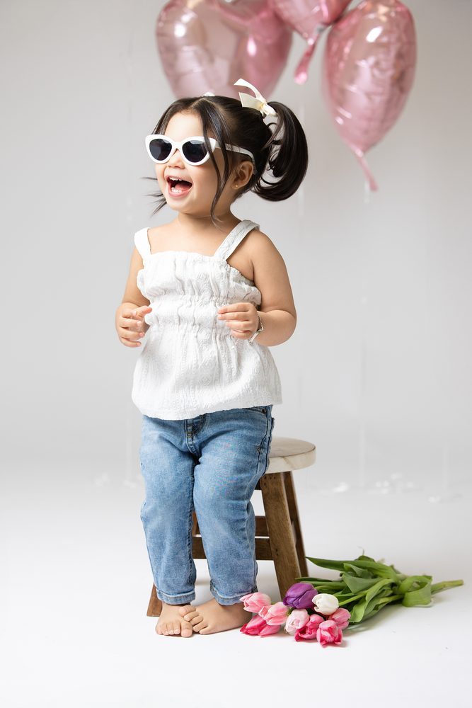 Laughing toddler girl in white sunglasses standing by a wooden stool, pink heart balloons and tulips nearby