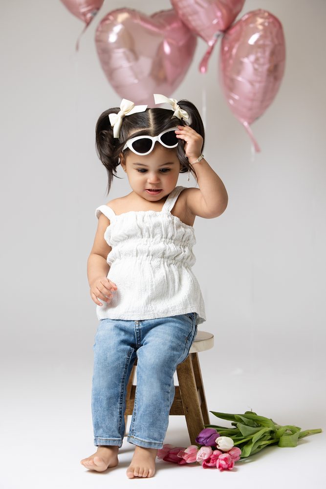 Toddler girl adjusting her white sunglasses on a wooden stool, pink and purple tulips at her feet
