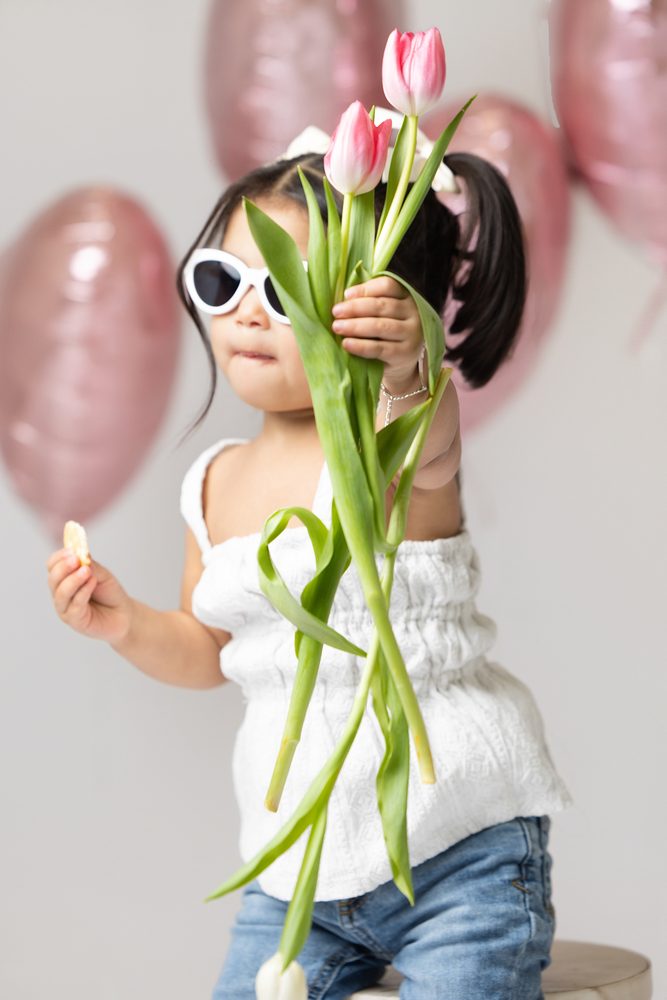 Toddler girl in white sunglasses holding pink tulips toward the camera in front of pink heart balloons