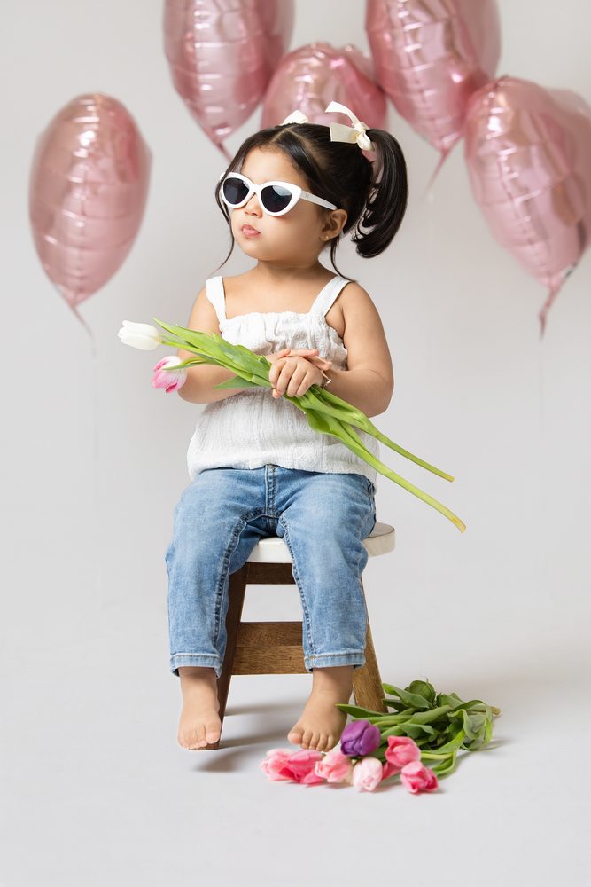 Toddler girl in sunglasses seated on a wooden stool cradling tulips, pink heart balloons floating behind