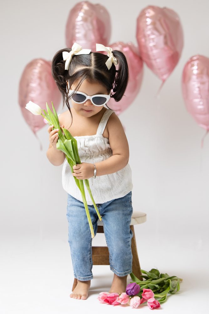 Little girl with pigtail bows looking down at a bunch of white tulips, pink heart balloons behind her