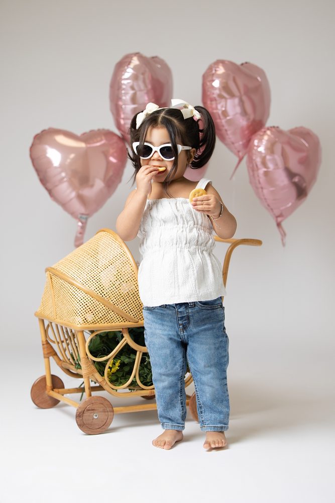 Girl in white sunglasses nibbling a cookie beside a rattan doll pram, pink heart balloons overhead