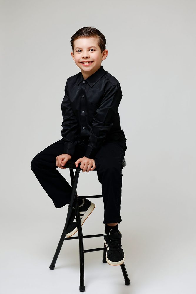 Smiling boy in an all-black outfit sitting on a tall black stool against a light gray backdrop