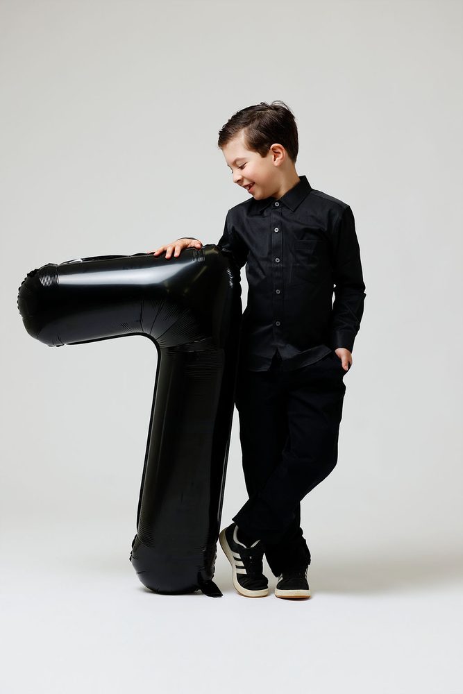 Boy in a black shirt leaning on a giant black '7' balloon with a hand in his pocket, smiling down