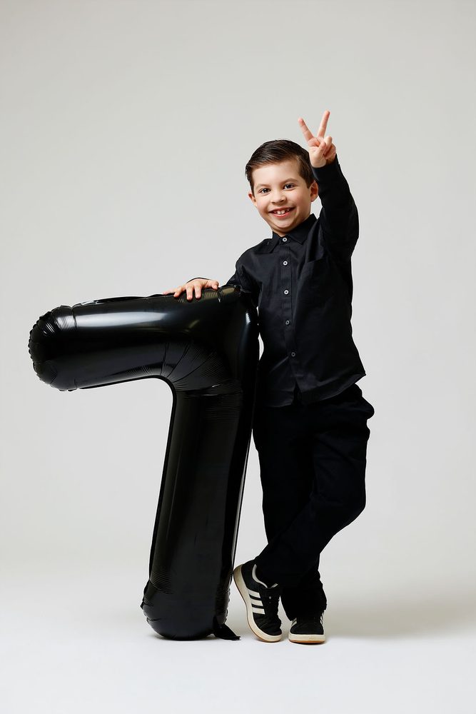 Boy in black flashing a peace sign with one hand resting on a giant black '7' balloon