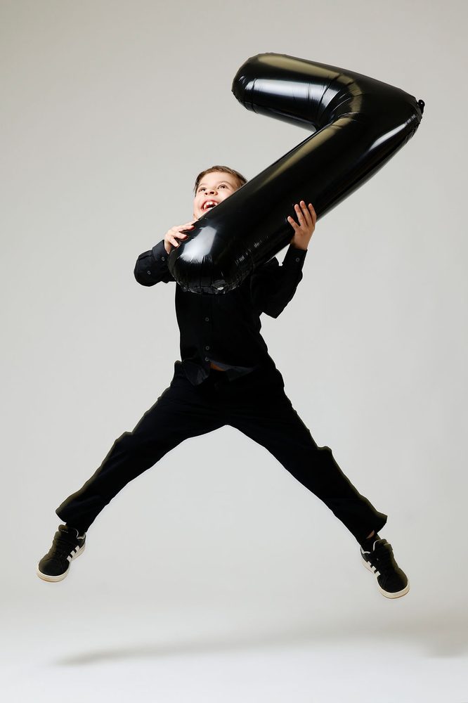 Boy mid-jump with legs spread wide, laughing and hugging a black '7' balloon in the air