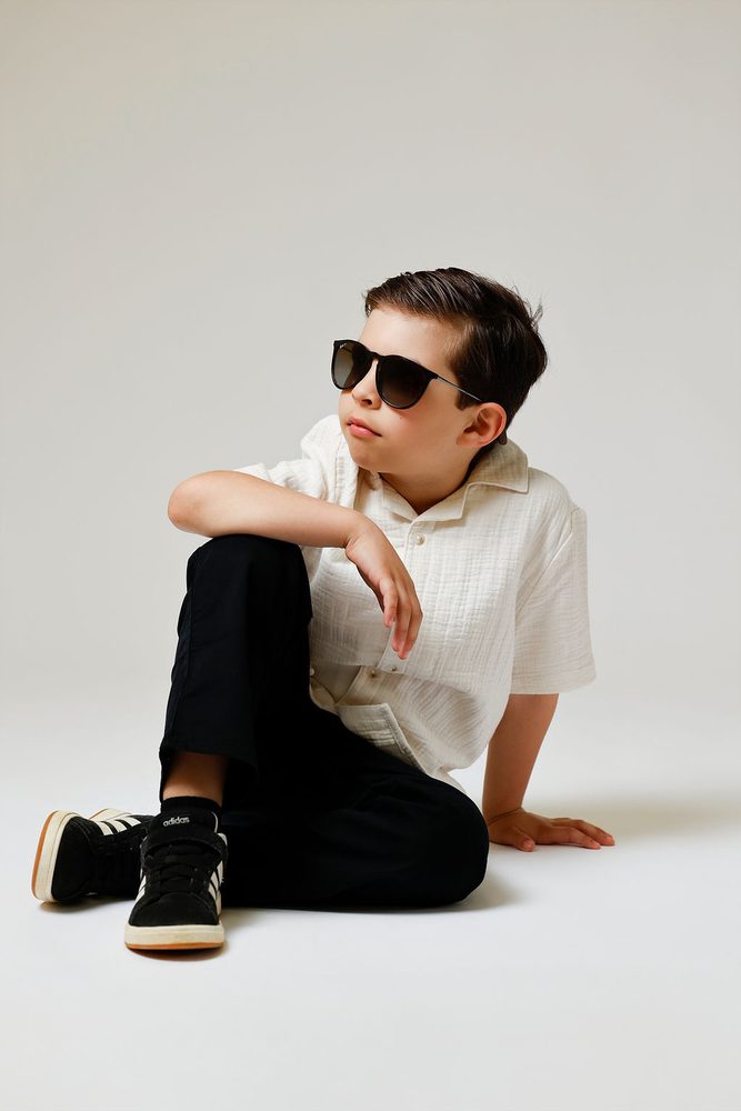 Boy in dark sunglasses and a cream linen shirt sitting on the studio floor, glancing to the side