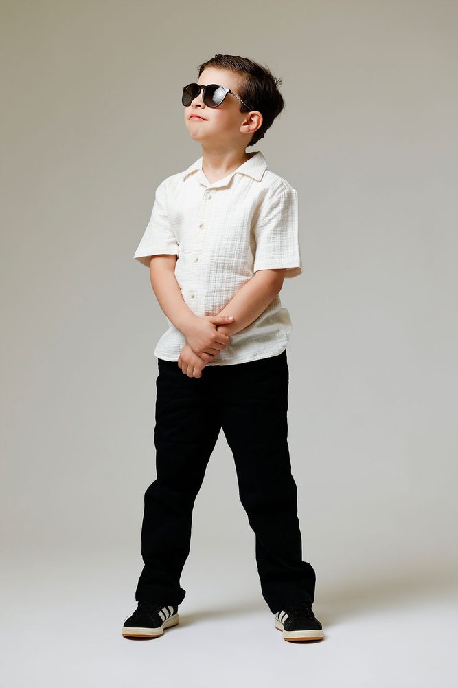 Boy in sunglasses standing with clasped hands and chin up, wearing a cream shirt and black pants
