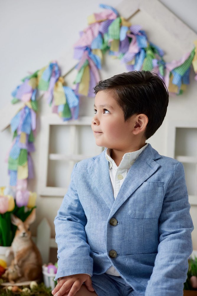 Boy in a blue linen blazer gazing sideways in front of a pastel ribbon garland and toy bunny