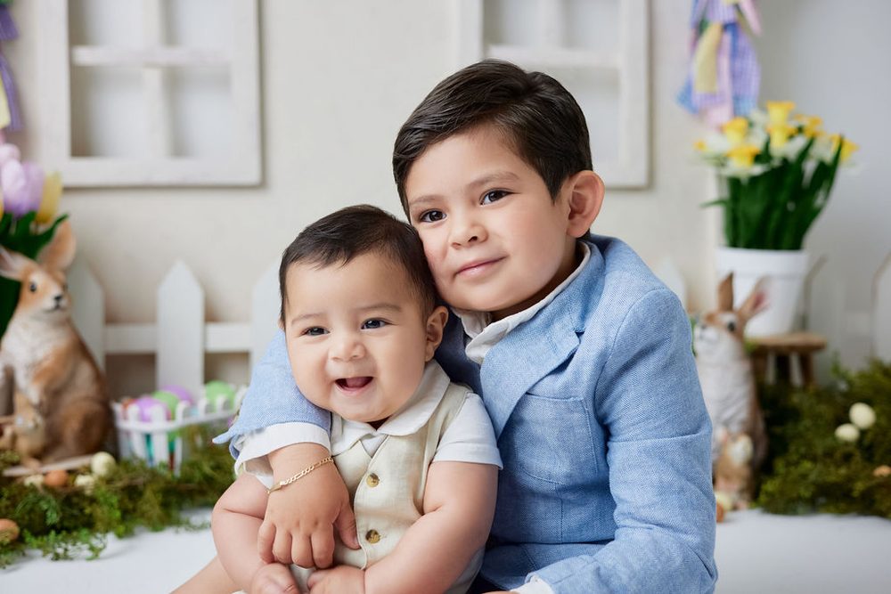 Older brother in a blue blazer hugging his smiling baby brother in a spring set with bunnies and daffodils