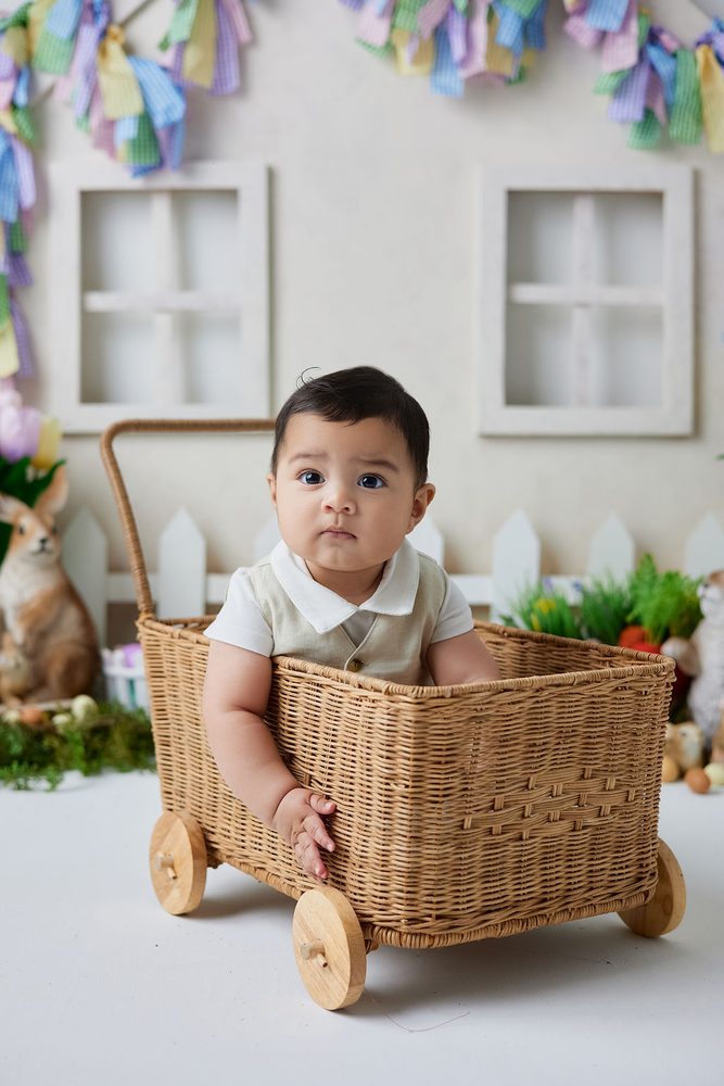 Baby in a cream vest sitting in a wicker wagon with wooden wheels in front of a spring cottage backdrop
