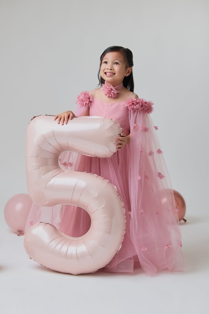 Girl in a pink tulle gown beaming upward while holding a large blush '5' balloon
