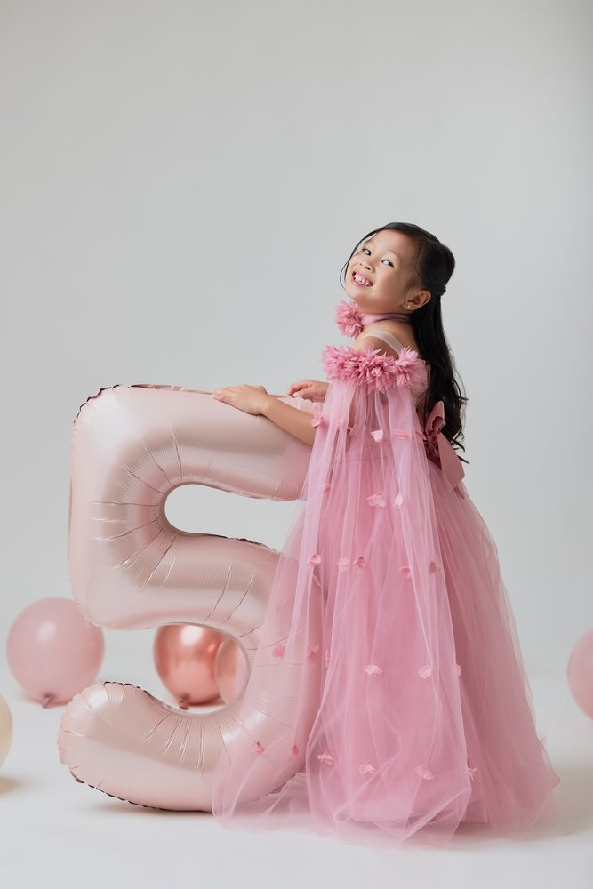 Girl in a pink gown leaning on a blush '5' balloon and smiling back over her shoulder