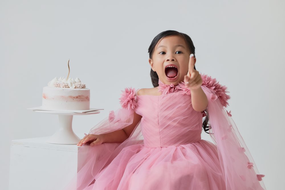 Excited girl in a pink dress pointing a frosting-tipped finger beside a white cake on a pedestal stand
