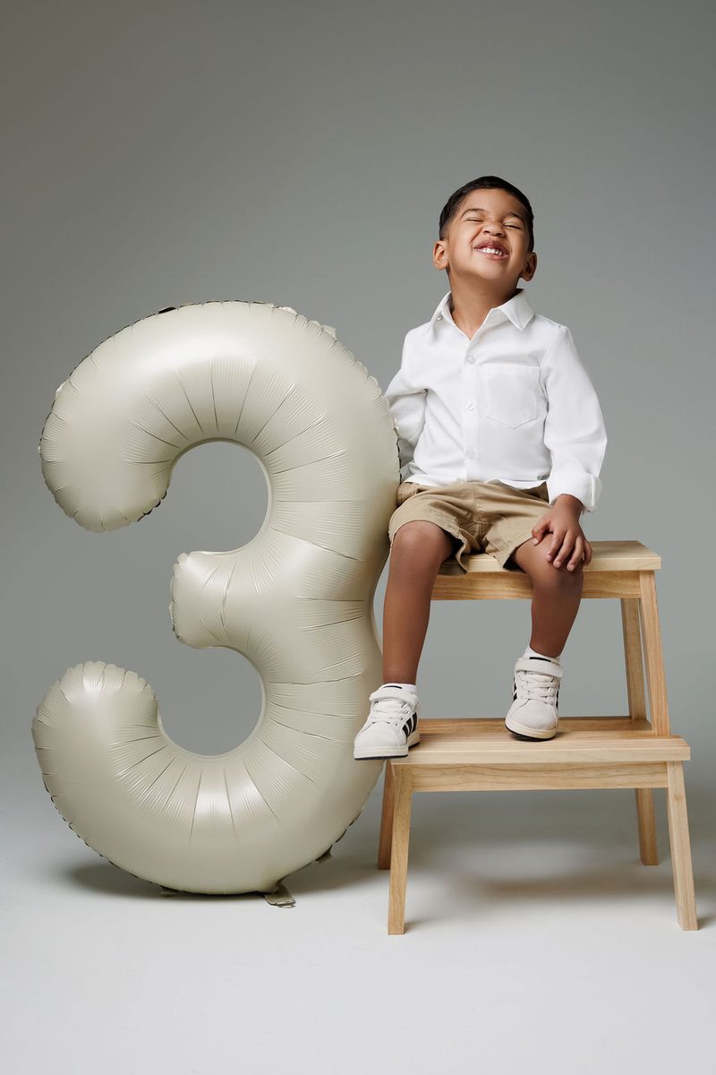 Kids birthday photography: laughing little boy on a wooden step stool beside a giant cream '3' balloon