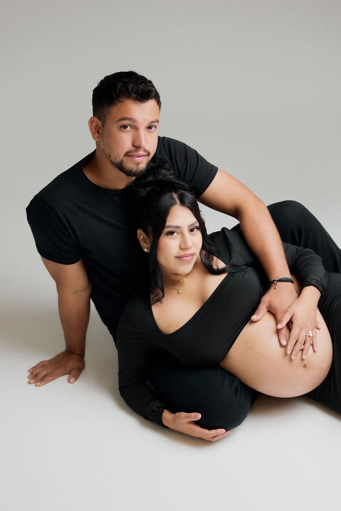 Expectant couple in black lounging on a white studio floor, his hand resting on her bare belly