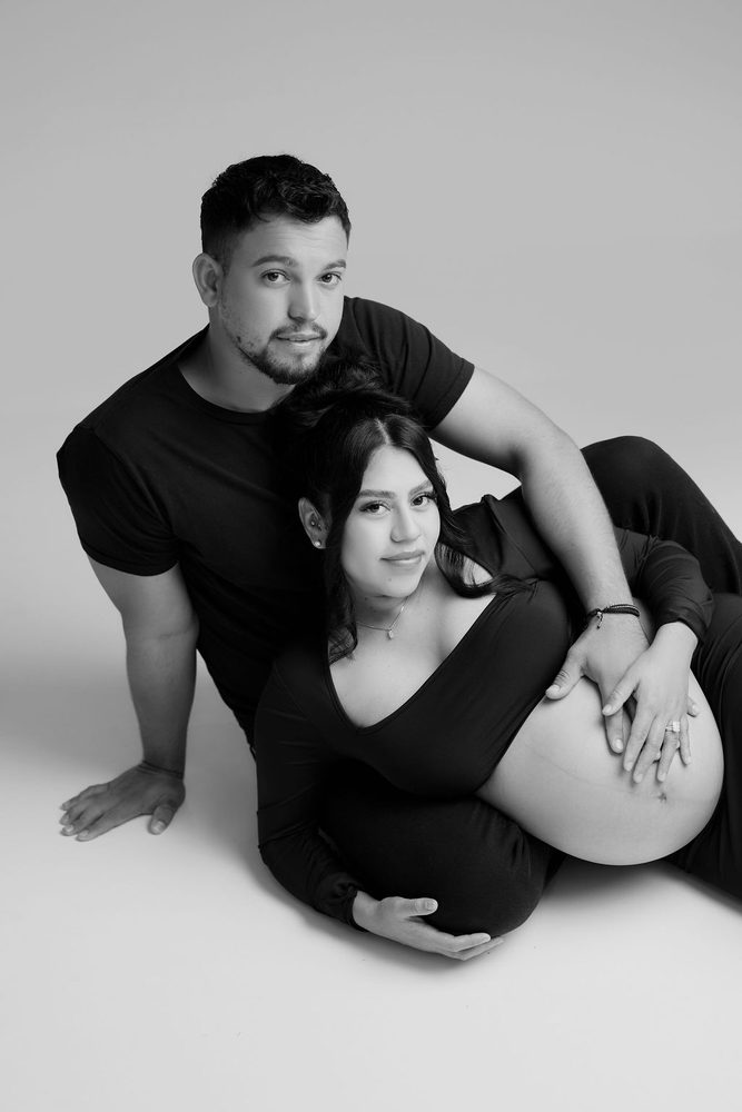Black-and-white studio portrait of an expectant couple reclining together, their hands on her belly