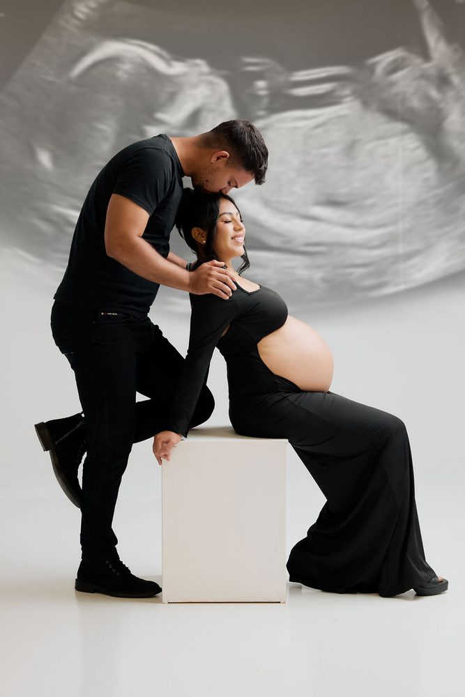 Partner kisses the expectant mother's forehead as she sits on a white block before a projected ultrasound image