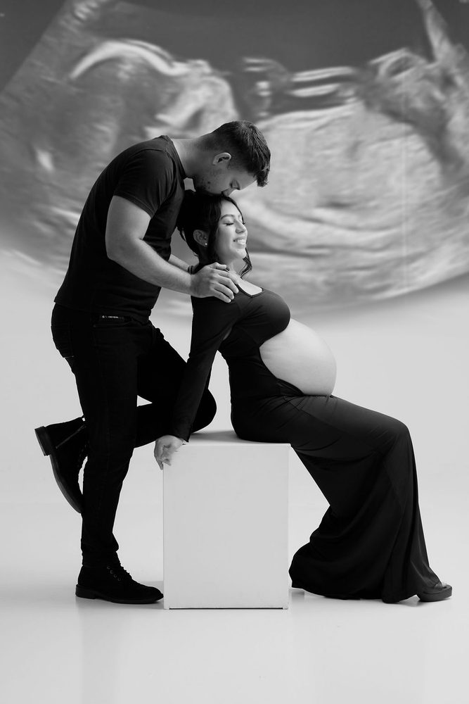 Black-and-white forehead kiss in front of a large ultrasound projection, the mother seated on a studio block
