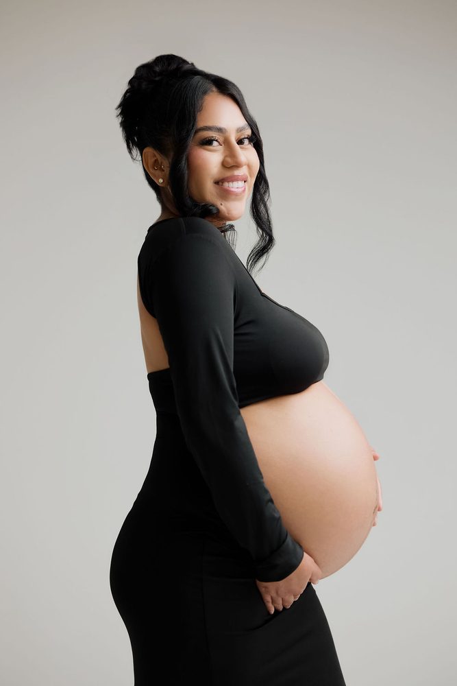 Smiling expectant mother in a black open-back crop top, shown in profile against a gray studio backdrop