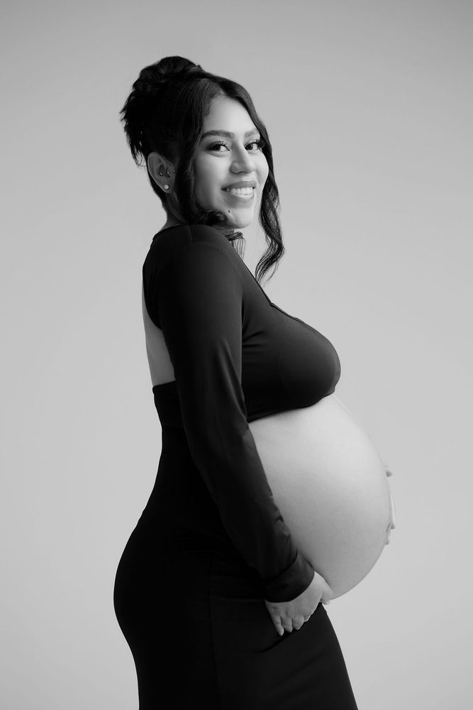 Black-and-white profile of a smiling expectant mother in a backless crop top, hands cradling her bare belly