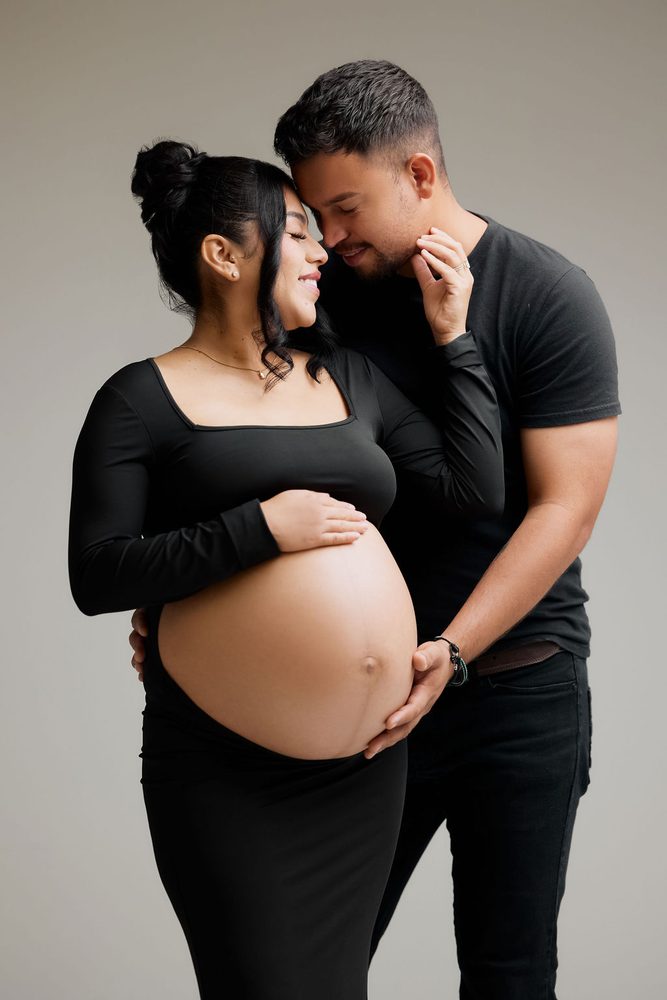 Expectant couple nuzzling in the studio, her hand on her bump while his hand supports it from below