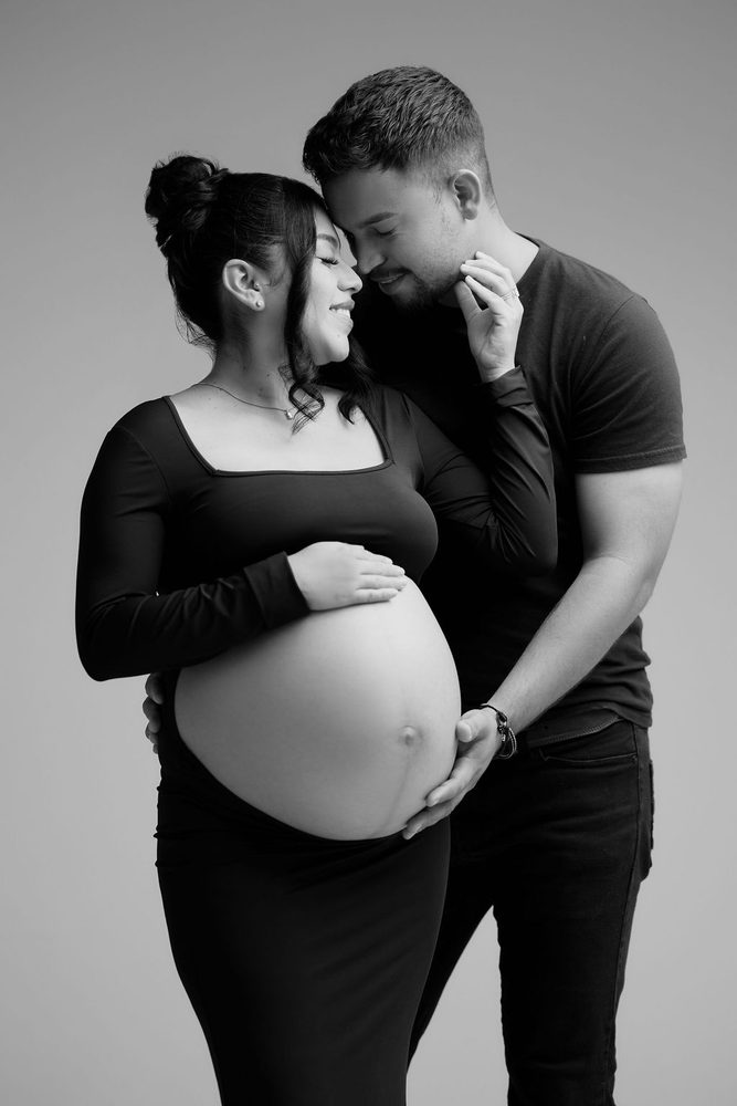 Black-and-white tender moment between expectant parents, foreheads close and his hand beneath her bare belly