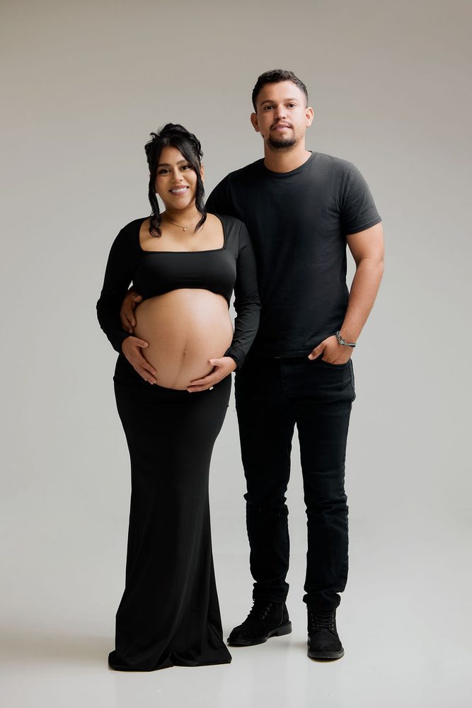 Full-length color studio portrait of an expectant couple in black, her hands framing her bare bump