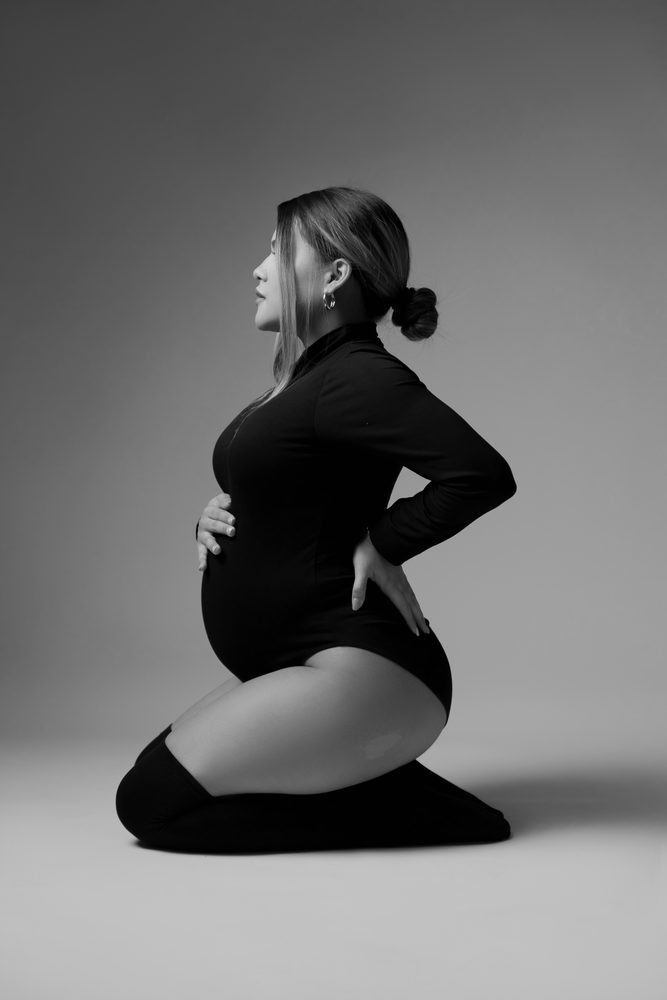 Black-and-white profile of an expectant mother kneeling in a turtleneck bodysuit, one hand on her belly