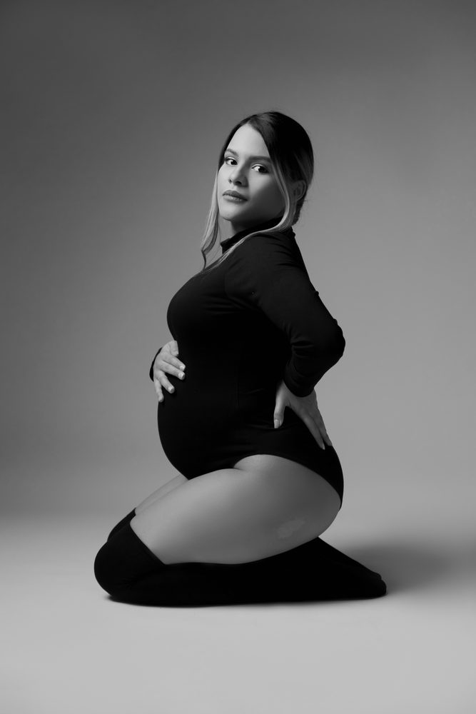Black-and-white kneeling pose, the expectant mother glancing over her shoulder in a black bodysuit
