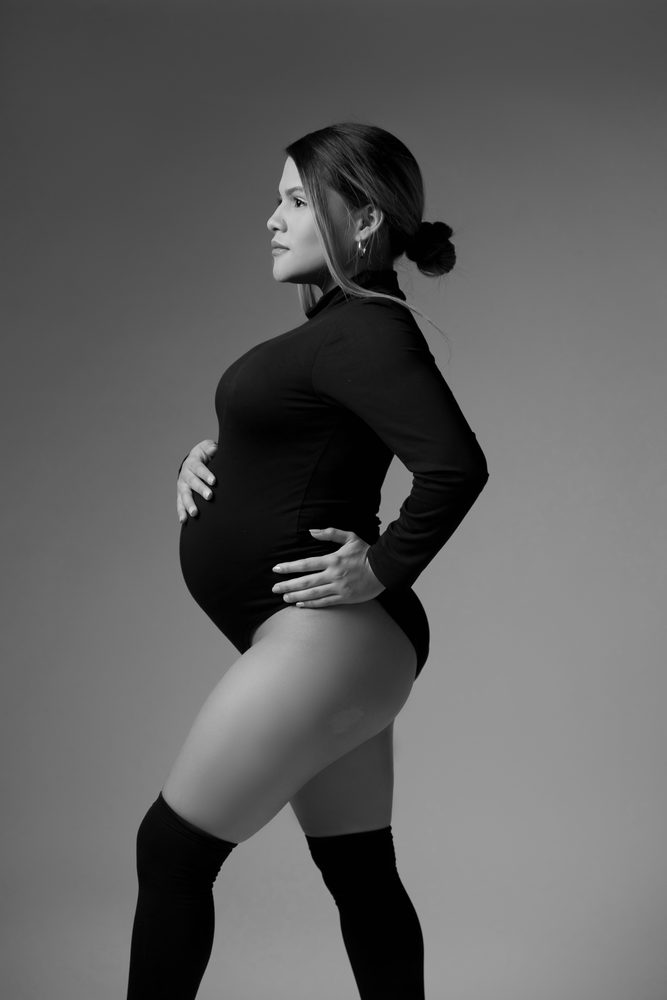 Black-and-white standing profile of an expectant mother in a turtleneck bodysuit, one hand on her bump