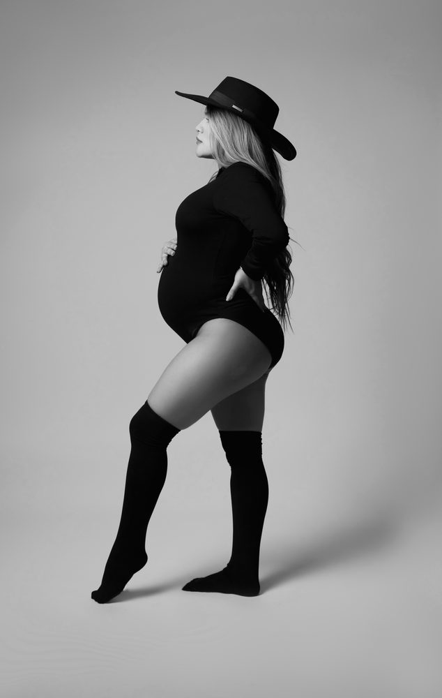 Black-and-white full-length pose of an expectant mother in a wide-brim hat, bodysuit and thigh-high socks