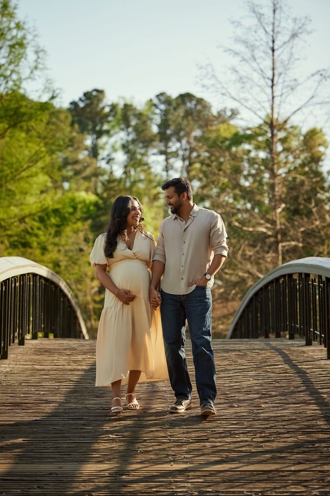 Expectant couple walking hand in hand across a wooden footbridge in a sunny wooded park