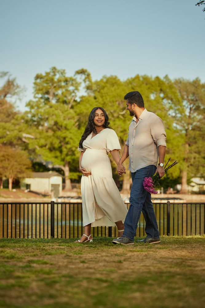 Expectant parents strolling on the grass beside a lakeside fence, her partner carrying pink flowers