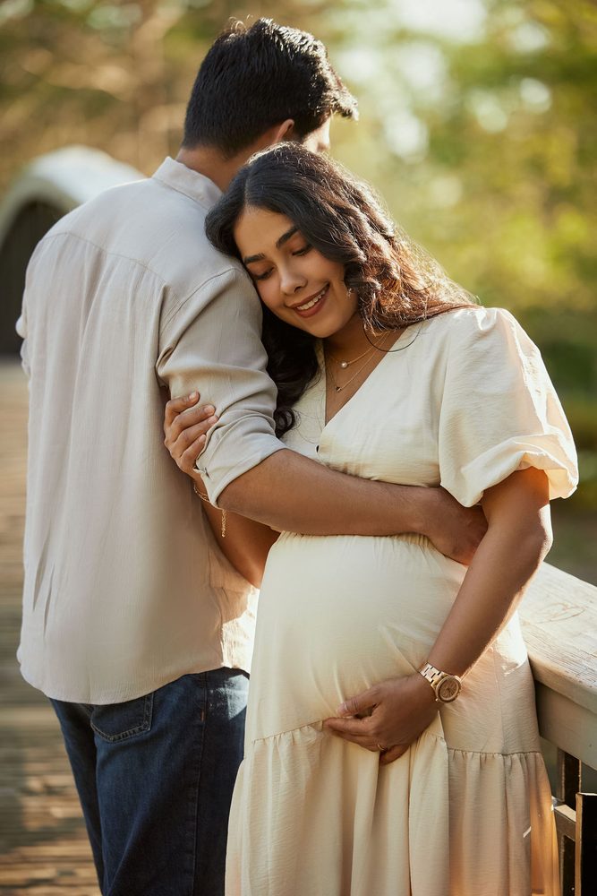 Expectant mother resting her head on her partner's shoulder on a footbridge, one hand on her belly