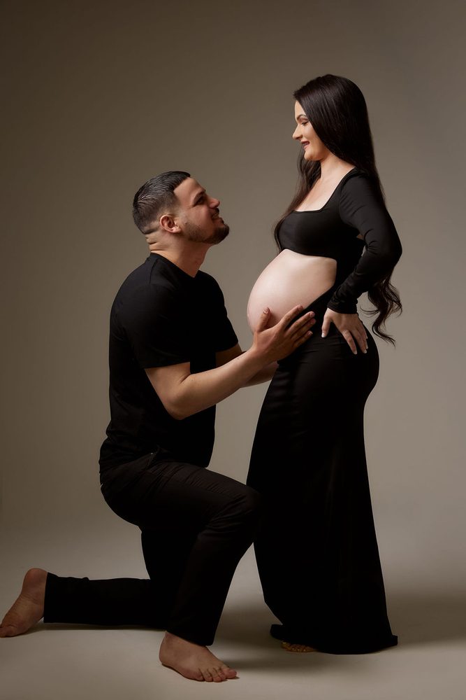 Partner kneels with his hands on the expectant mother's bare belly as she smiles down at him in the studio