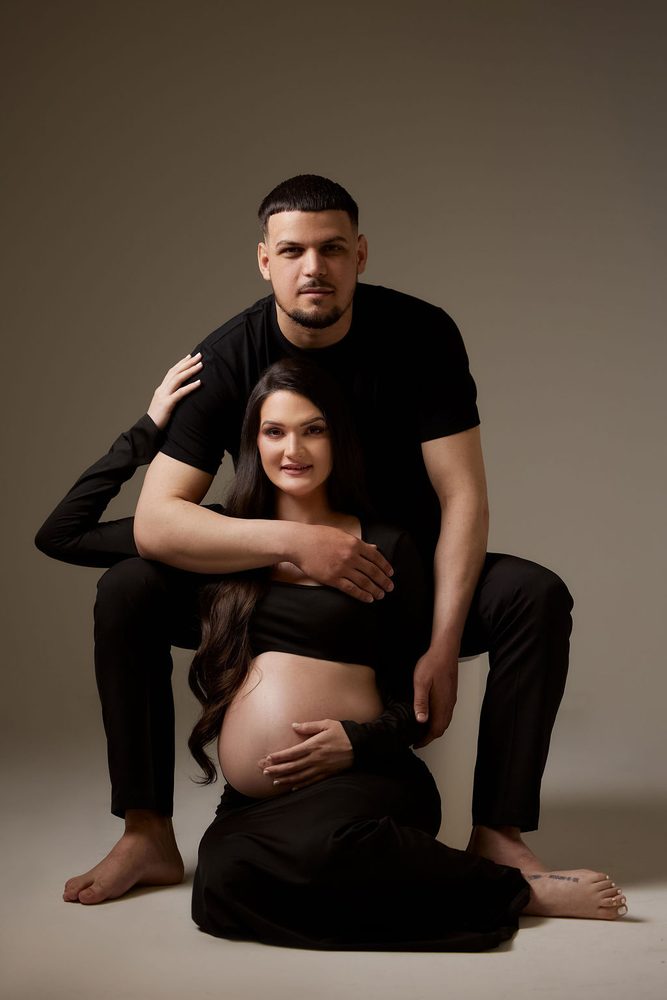 Expectant mother seated on the floor between her partner's knees, his arm across her, warm gray backdrop